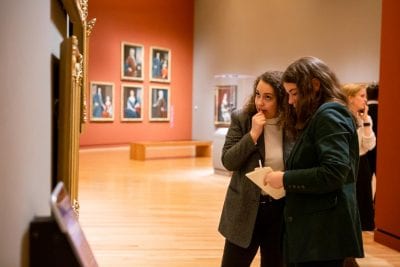 Two young women study paintings in a museum gallery.