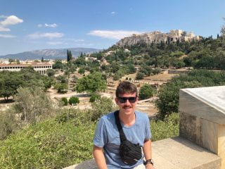 Image of the author in a blue shirt seated in front of Greek Ruins. 