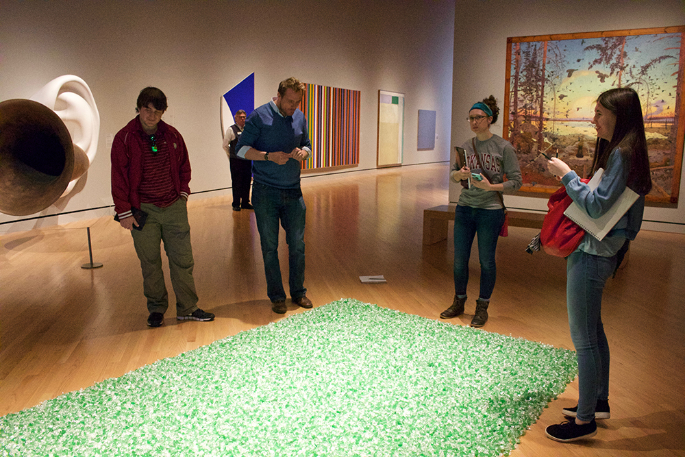 Professor and students examine a work of art consisting of a large square on floor filled in with candy. 